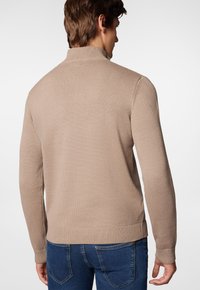 Beżowy sweter dzianinowy z kołnierzem stojącym, ściągaczami przy mankietach i u dołu. Charakteryzuje się teksturowanym materiałem i luźnym krojem, zestawiony z ciemnymi dżinsami.