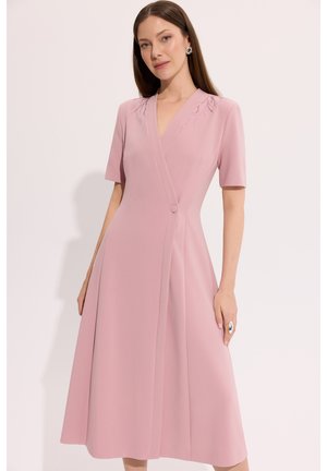 Kobieta ubrana w różową sukienkę do kolan z krótkim rękawem, o kroju kopertowym i subtelnymi zdobieniami przy dekolcie, stojąca na białym tle.