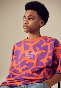 Oranje en paarse T-shirt met korte mouwen en abstract patroon. Heeft een grafisch ontwerp met de tekst "LOS ANGELES CALIFORNIA SINDS 1982".