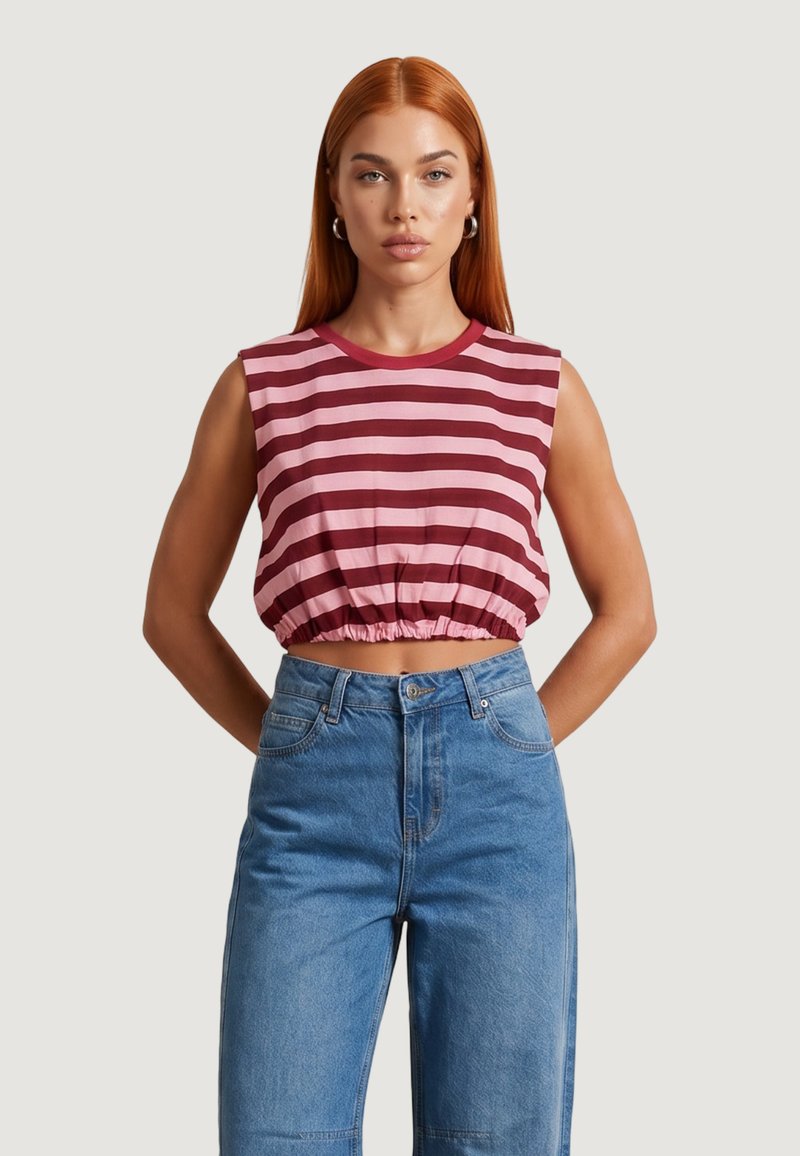 Mujer con cabello largo y rojo, que lleva una camiseta corta sin mangas a rayas rosas y rojas y unos vaqueros azules de talle alto, de pie con las manos detrás de la espalda.