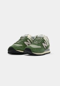 Žalios ir smėlinės New Balance 574 sportbačiai su pilkomis raištelėmis ir juodais padais ant balto fono.