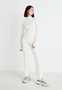 Completo athleisure bianco attillato composto da giacca con zip e colletto alto e pantaloni a zampa, abbinato a sneaker grigie e bianche.