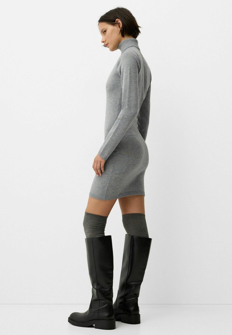 Frau in einem enganliegenden grauen Rollkragenkleid, mit knielangen grauen Socken und schwarzen kniehohen Stiefeln, die im Profil vor einem weißen Hintergrund steht.