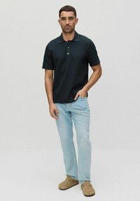 Svart polo med krage och tre knappar, tillsammans med ljusblå jeans och beigea slip-on skor, stående mot en ljus bakgrund.