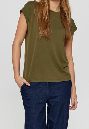 Kobieta ubrana w oliwkowozieloną koszulkę z krótkim rękawem, lekko włożoną do ciemnoniebieskich spodni, z jedną ręką w kieszeni na jednolitym tle.