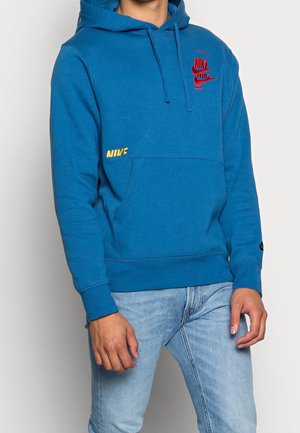 Mann trägt einen blauen Nike-Hoodie mit roten und gelben Logoaufdrucken, kombiniert mit hellblauen Jeans vor einem schlichten weißen Hintergrund.
