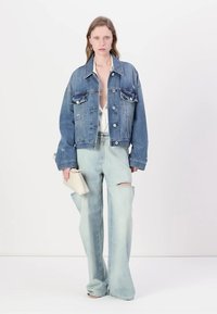 Veste en denim bleu moyen avec poches poitrine, associée à un jean large bleu clair avec des fentes au genou. Le modèle tient un petit sac beige.
