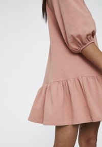 Rosa Baumwollkleid mit Puffärmeln und gerafftem Rüschensaum, das eine lockere Passform und eine sanfte Textur aufweist. Sichtbare Nahtdetails an der Taille.