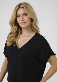 Haut noir à col en V avec des manches courtes, présentant des accents froncés. Tissu doux et léger avec une texture lisse. Design simple et polyvalent.