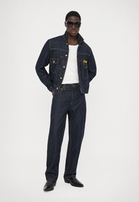 Chaqueta de mezclilla oscura con cuatro bolsillos y bordado amarillo en el pecho, combinada con pantalones de mezclilla oscura a juego y botas vaqueras negras.