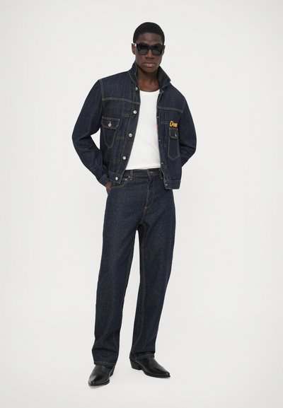 Mørk denimjakke med fire lommer og gul broderi på brystet, parret med matchende mørke denimbukser og sorte cowboystøvler.