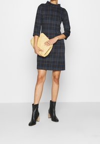 Laivastonsininen ruudullinen mekko korkealla kauluksella ja kolmannen neljänneksen hihoilla. Malli pitää keltaista laukkuansa. Mustat nilkkurit suppilomaalla korolla.