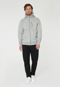 Sudadera gris con cremallera y capucha con cordón, que presenta un detalle de logo negro. Combinada con pantalones negros y zapatillas blancas, todo sobre un fondo sencillo.