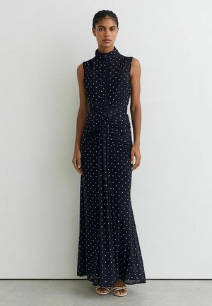 Frau steht vor einer weißen Wand und trägt ein ärmelloses schwarzes Maxikleid mit weißen Punkten und weiße Sandalen.