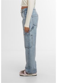 Gai zila denima kravas bikses ar brīvu piegriezumu, kurām ir lielas sānu kabatas un šūšanas detaļas, valkā ar rozā un brūnām kedām.