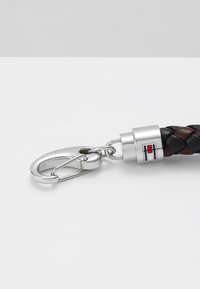 Breloacă din piele împletită, de culoare neagră și maro, cu un sistem de închidere din metal argintiu. Include un conector cilindric argintiu cu un accent de logo.
