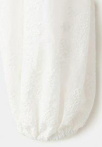 Tissu blanc avec une broderie florale subtile et un poignet élastique froncé en bas, créant une apparence douce et texturée.