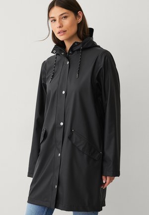 Kobieta w czarnej, zapinanej na zatrzaski przeciwdeszczowej pelerynie z kapturem i dwoma ukośnymi kieszeniami z patkami, stojąca na tle jednolitego jasnego tła.