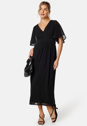 Robe maxi noire en tissu léger avec décolleté en V, manches flottantes et taille élastique. Associée à un sac à main noir texturé et des sandales.