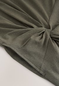 Olivgrüner Stoff, in der Mitte gedreht, zeigt feine Strickstruktur und eingefasste Kanten auf einer hellen Oberfläche.