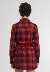 Rochie camasă în carouri roșu și negru, realizată dintr-un material moale, cu mâneci lungi, manșete cu butoane și o curea la talie. Vedere din spate, cu un tiv curbat.