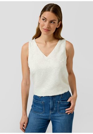 Frau mit hellbraunen Haaren, die ein weißes, ärmelloses, strukturiertes Top mit V-Ausschnitt und blaue, hoch taillierte Jeans mit Vorder taschen trägt und zur Seite blickt.