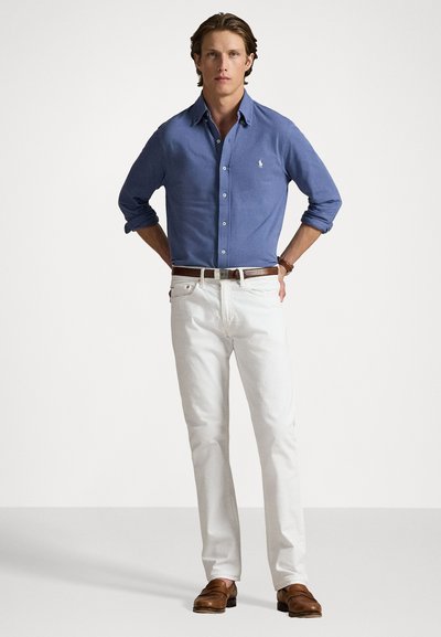 Camicia blu con bottoni, pantaloni bianchi, cintura marrone e mocassini marroni. Presenta un piccolo logo sulla camicia. Tessuto liscio e design aderente.