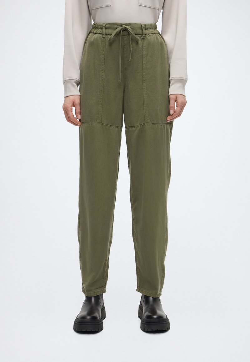 Άτομο που φοράει ελιά πράσινο παντελόνι με κορδόνι στη μέση και εμφανείς ραφές, μαζί με μαύρα στιβαρά μποτάκια, στέκεται μπροστά σε ένα απλό ανοιχτόχρωμο φόντο.