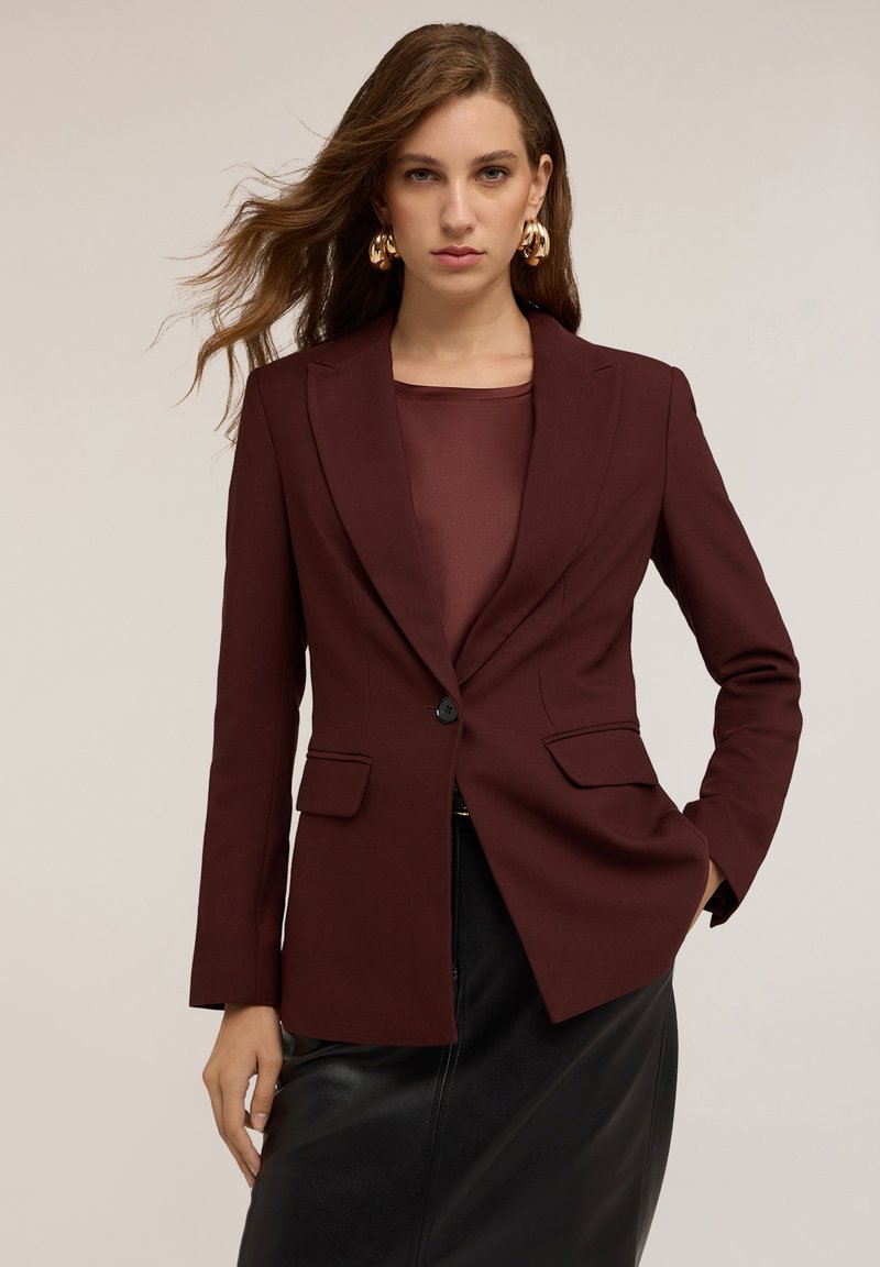 Donna con capelli mossi che indossa orecchini a cerchio d'oro, un blazer bordeaux sopra un top coordinato e una gonna di pelle nera, su uno sfondo neutro.