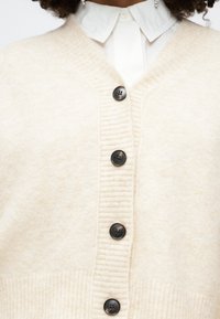 Bežový vlněný svetr s V-výstřihem, ribbovanou strukturou, šesti černými knoflíky a jemným povrchem, navrstvený na bílé košilové tričko.