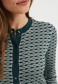 Stickad cardigan med ett turkos och ljusgrått vågmönster, med rund hals, rad av svarta knappar och textilstruktur.