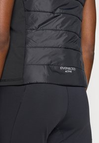 Veste noire matelassée avec des panneaux latéraux en tissu extensible, présentant des sections matelassées horizontales, et un logo sur l'ourlet inférieur.