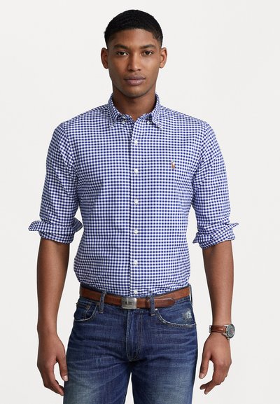 Chemise en vichy bleu et blanc avec col boutonné, manches retroussées et un petit logo. Portée avec un jean foncé et une ceinture marron.