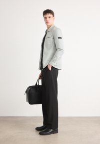 Ανοιχτό πράσινο ριγέ πουκάμισο, μαύρο παντελόνι και μαύρα παπούτσια. Το μοντέλο κρατά μια μαύρη τσάντα. Το ρούχο έχει στενή εφαρμογή και λεπτομέρεια ώμου.