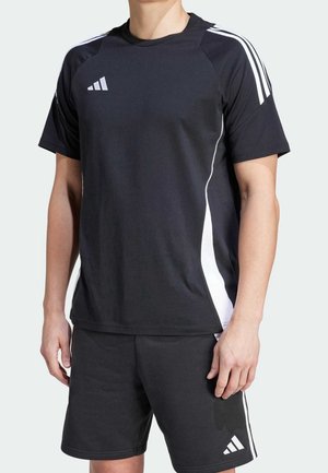 Mann trägt schwarzes Sportshirt und Shorts mit weißen Streifen und Logos, steht mit locker hängenden Armen an den Seiten vor hellem Hintergrund.