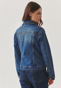 Jean jacket in keskipesty sinisessä, jossa on klassinen kaulus, napit ja kontrastivärinen ommel; sisältää hyvin istuvan muodon ja takakiinnityksen yksityiskohdan.