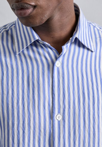 Camisa a rayas verticales azules y blancas con cuello puntiagudo, detalles de botones y un tejido de algodón texturizado.