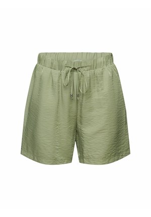 Femme portant un short à cordon vert clair, un haut court blanc et des sandales beiges à enfiler, debout avec une main sur la hanche.