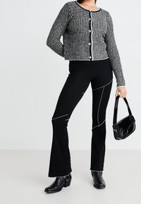 Cardigan tricoté noir et blanc avec boutons à l'avant, associé à un pantalon noir évasé avec des coutures blanches, et un petit sac à main noir.