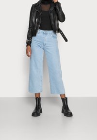 Giacca da motociclista in pelle nera sopra un top nero trasparente, abbinata a jeans cropped blu chiaro e ankle boots neri.