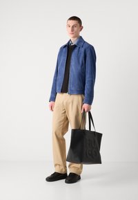 Μπλε σουέτ τζάκετ, μαύρο τοπ, χακί παντελόνι, και μαύρα αθλητικά παπούτσια. Μαύρη τσάντα tote με ανάγλυφο λογότυπο "HUGO". Το μοντέλο κοιτάζει μπροστά.