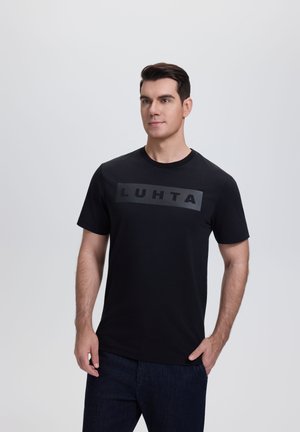 Mann trägt ein schwarzes T-Shirt mit dem grauen "LUHTA"-Logo, steht mit einer Hand in der Tasche vor einem einfarbigen hellen Hintergrund.