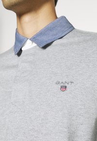 Harmaa polo-paita, jossa on sininen farkkukravatti, valkoinen nappi ja brodeerattu logo rinnassa. Pehmeä rakenne, rento muotoilu.
