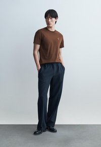 Jeune homme debout, les mains dans les poches, portant une chemise marron à manches courtes, un pantalon large noir et des chaussures noires, devant un fond uni.