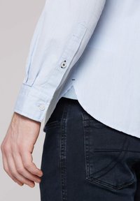 Helles blaues Button-Up-Hemd mit langen Ärmeln, das eine subtile Textur, einen abgerundeten Saum und Knöpfe an den Manschetten aufweist, kombiniert mit dunklen Jeans.