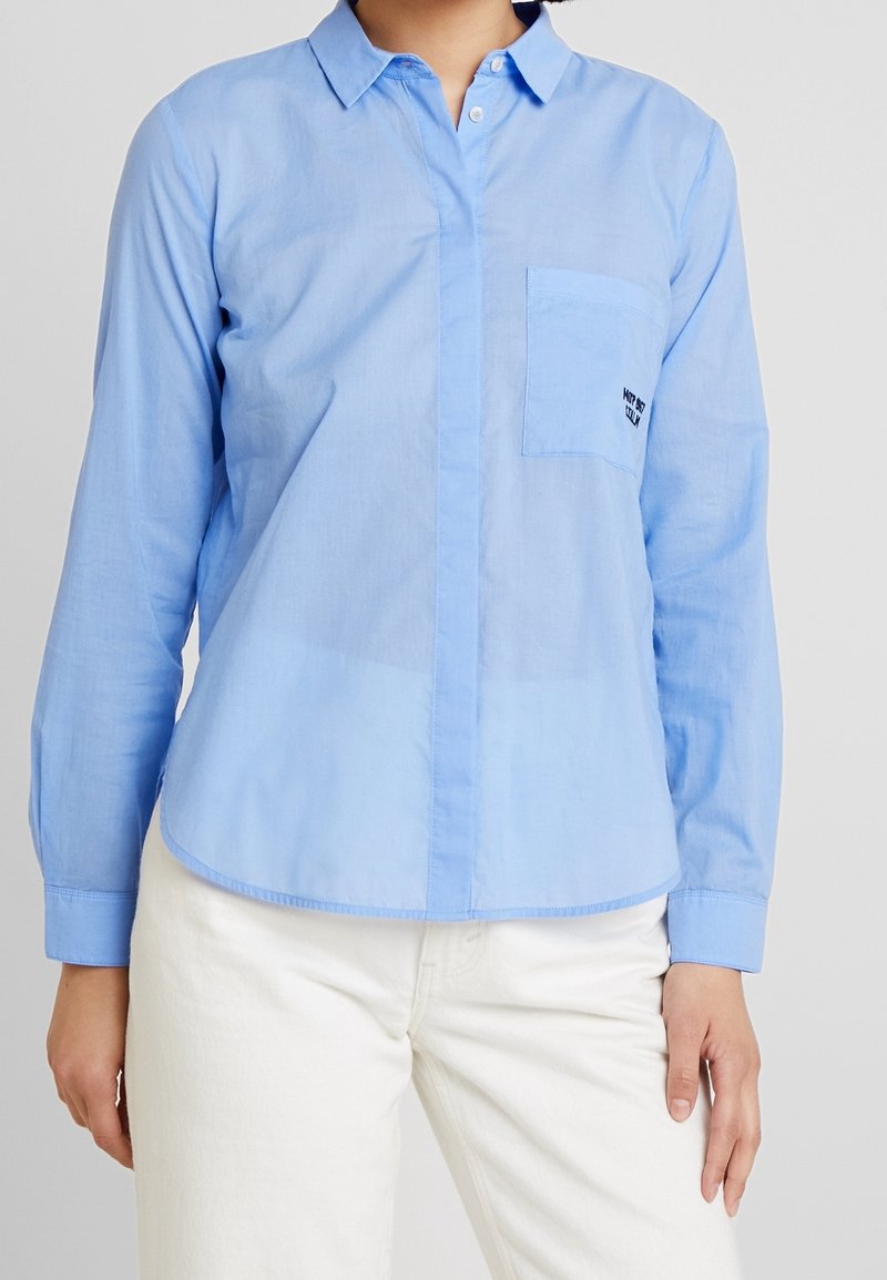 Camicia azzurra chiara in cotone, con maniche lunghe, taschino sul petto e orlo arrotondato. Ricamo semplice sulla tasca.