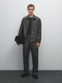 Giacca grigia con zip, maglione grigio a trama, pantaloni a pieghe abbinati e scarpe scure. Tiene una borsa in suede nera con tracolla.