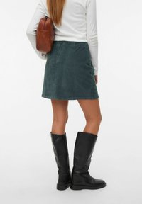 Jupe mini en velours côtelé vert avec une fermeture éclair dissimulée, associée à des bottes noires montant jusqu'aux genoux et à un sac en cuir marron, vue de dos.