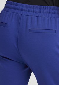 Tuvplāns zilas elastīgas jostas bikses ar divām aizmugurējām šķēlējamām kabatām, valkātas kopā ar melnu topu.