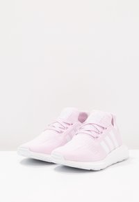 Pár světle růžových sportovních tenisek s bílými podrážkami a třemi bílými postranními pruhy, vystavených na bílé ploše s jednobarevným pozadím.